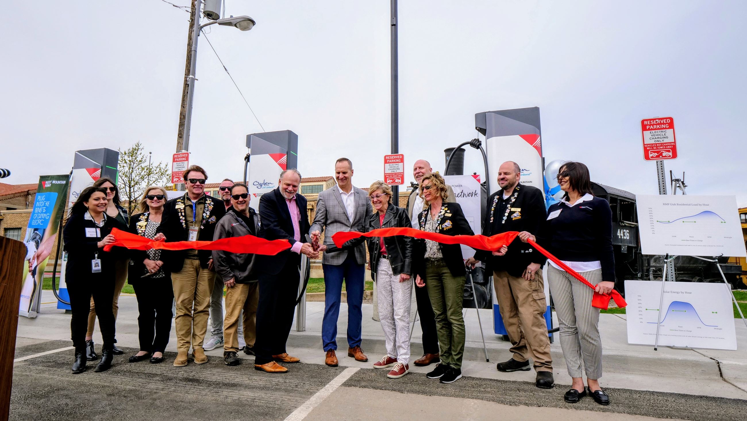 A group of people cutting a red ribbon with large scissors, in front of the new EV charging stations
